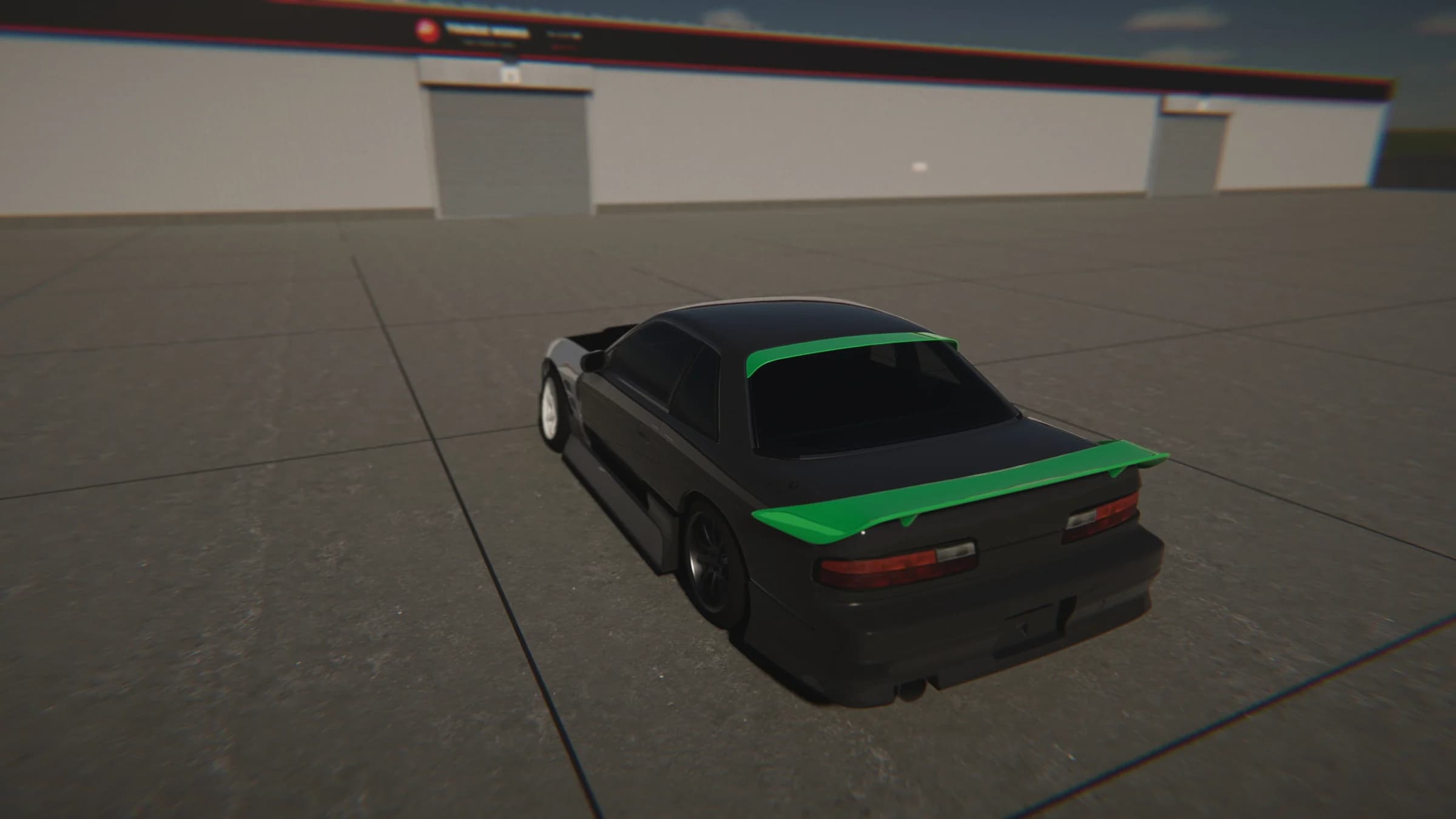 Dark coupe with green roof and rear-wing accents viewed from above in an open paved area beside garage buildings