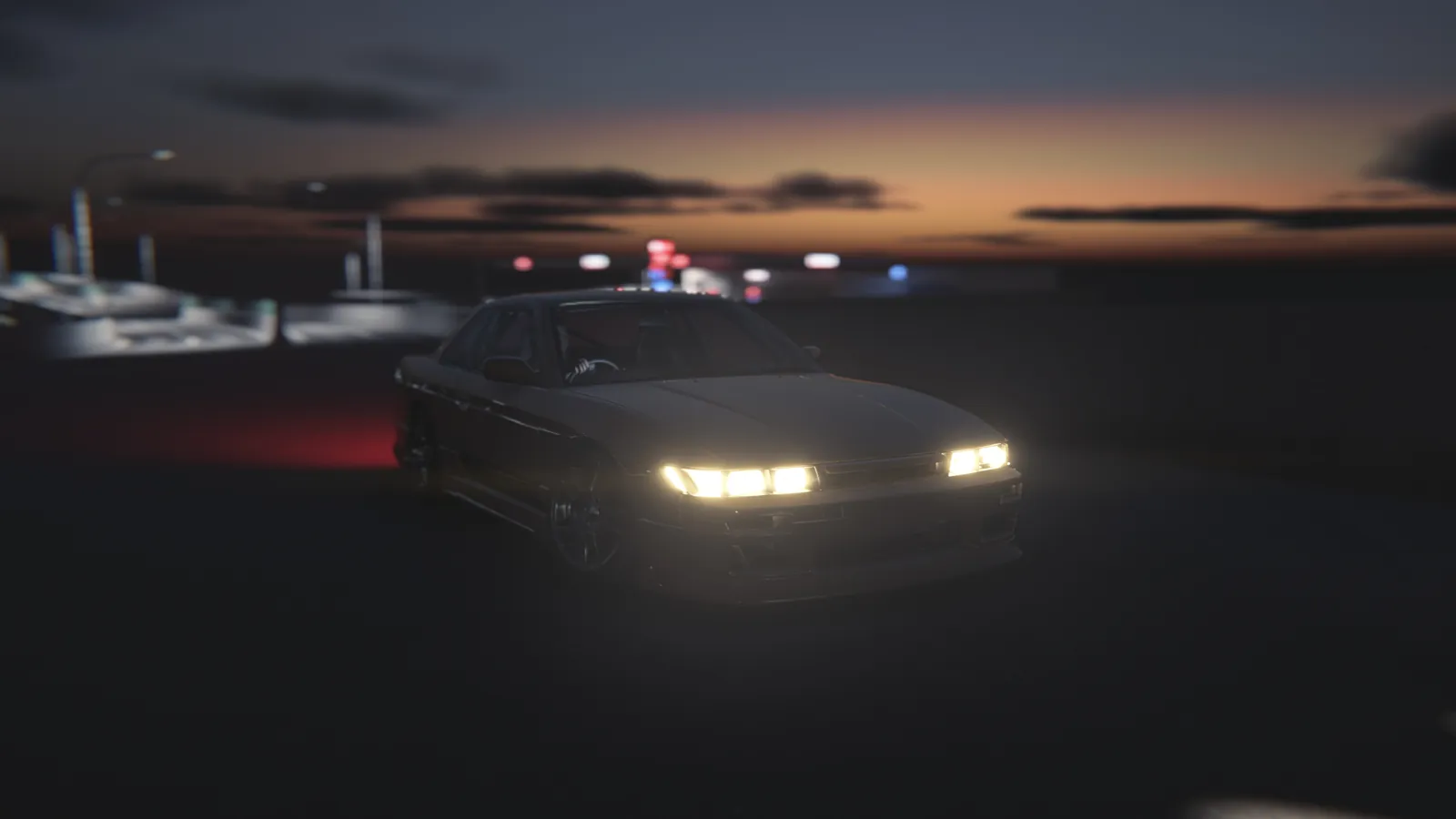 Coupe with headlights on facing the camera at dusk, with an orange sunset glow and the fuel station lights behind it